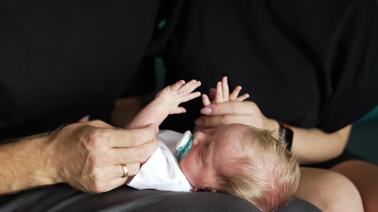 Sleeping newborn blond baby lies on a parent's laps. Mom and dad shaking tiny kid's hands but he continues to sleep. Close up.