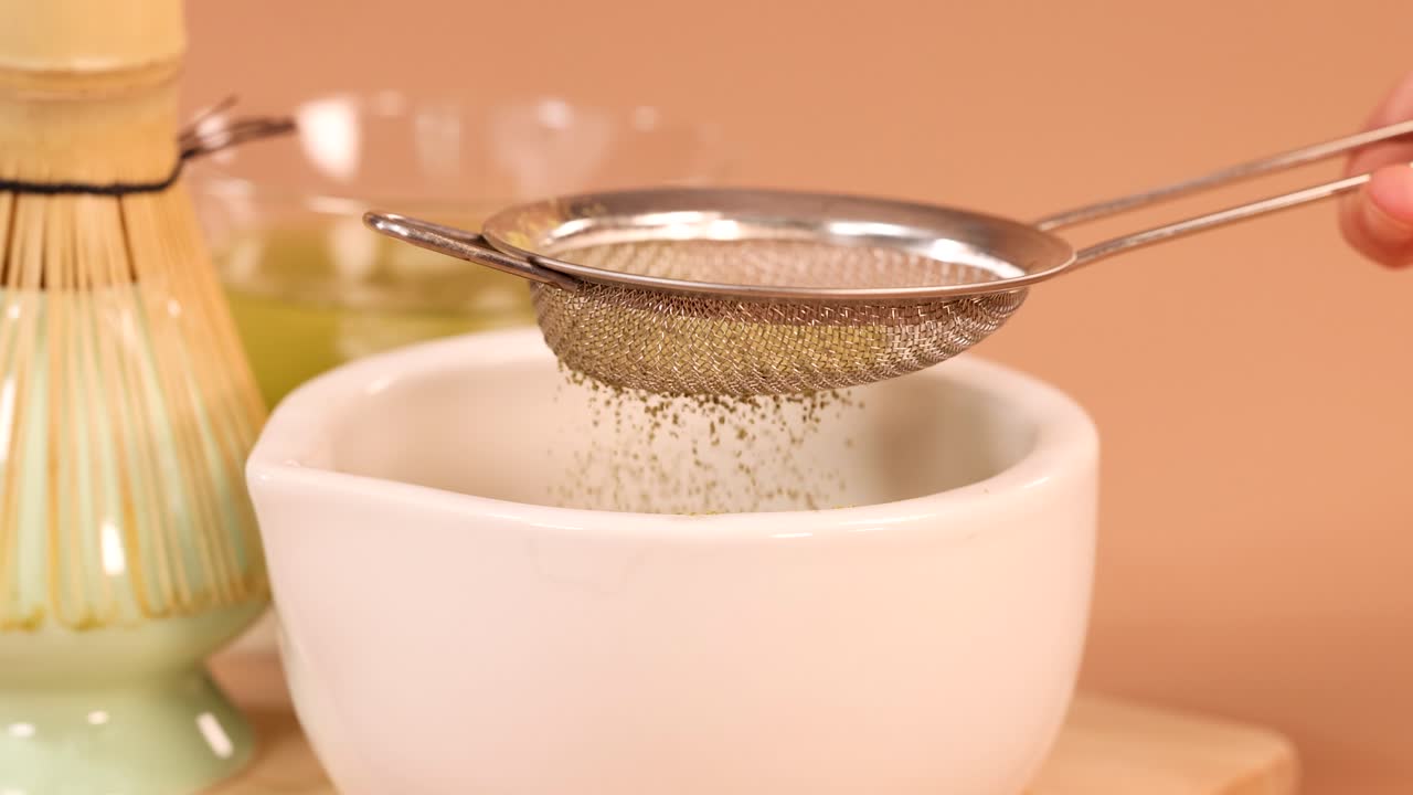 Hand sifts fine matcha powder through mesh strainer into bowl, soft lighting, traditional tea tools