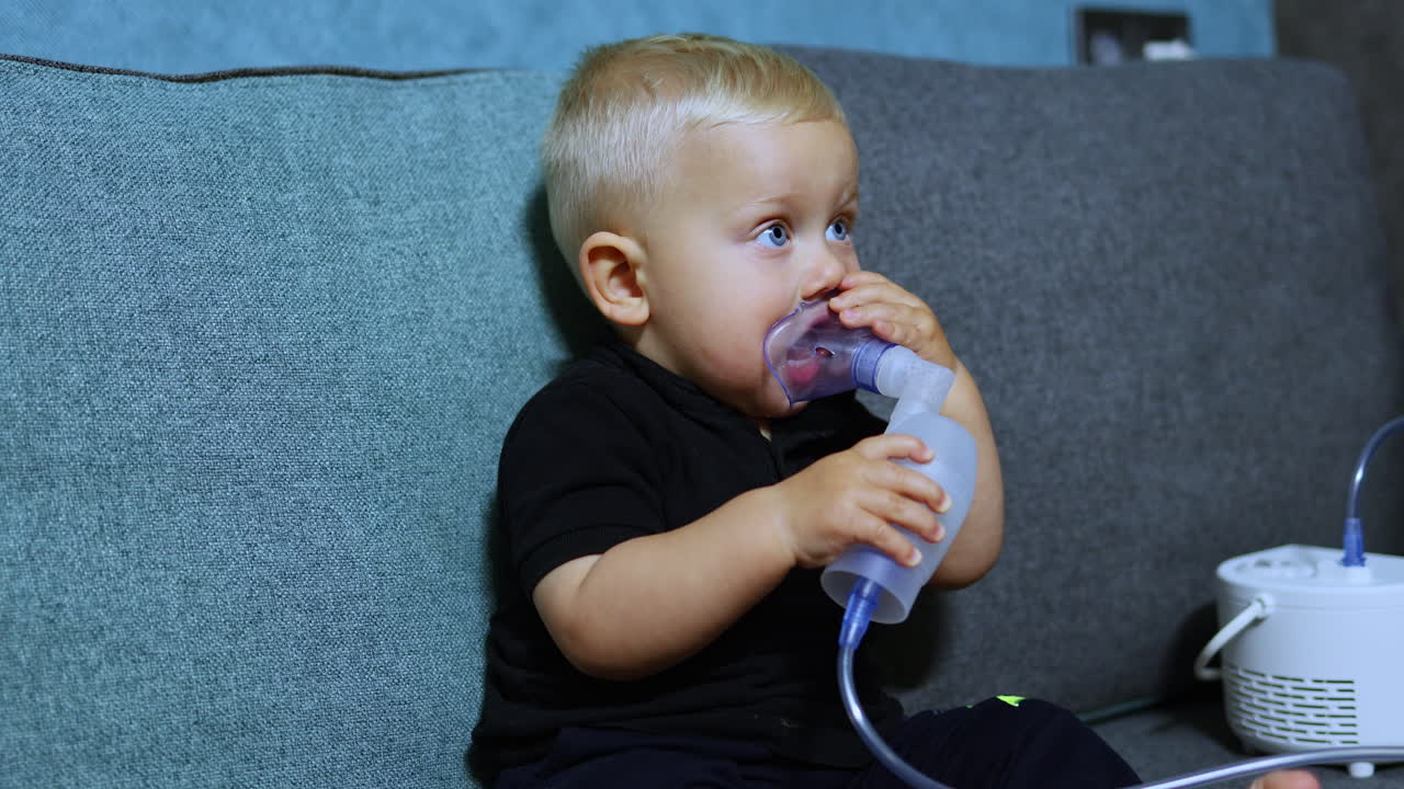 En liten pojke sitter i soffan och andas med en inhalator. Terapeutisk inhalation för barn. Inhalationsmask för barn