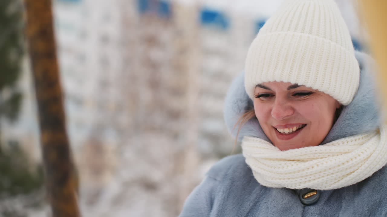 Mãe sorrindo em um balanço no parque de inverno, usando um casaco azul macio, gorro e cachecol de malha brancos. Retrato em close-up de uma mãe desfrutando de um parquinho nevado e tranquilo, com um sorriso gentil e a respiração visível, e prédios de apartamentos urbanos ao fundo.