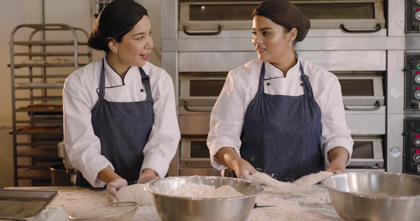 Diverse female bakers in chef coats scooping flour, kneading dough into proofing baskets at bakery