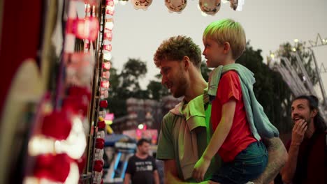 A-dad-with-curly-hair-in-a-Green-T-shirt-holds-his-little-blond-son-in-a-red-T-shirt-in-his-arms-and-they-stand-near-the-ticket-office-in-an-amusement-park-that-is-equipped-with-bright-light-bulbs