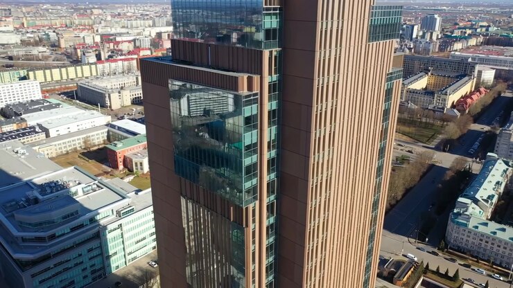 La telecamera scivola accanto a un grattacielo contemporaneo, mostrando linee geometriche della facciata, ampie sezioni di finestre riflettenti, edifici vicini, strade e un vasto paesaggio urbano in pieno giorno