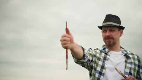 close-up of an artist wearing a hat and a checked shirt, holding a painting brush and pointing it to determine an angle. the artist is focused, capturing the perfect composition in an outdoor setting