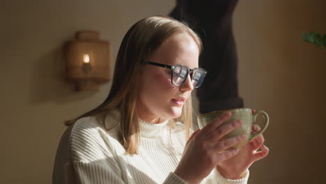 young woman in white sweater enjoys sweet coffee with soft smile, holding cup near face as sunlight gently highlights her expression, seated indoors beside decorative lamp and wall art