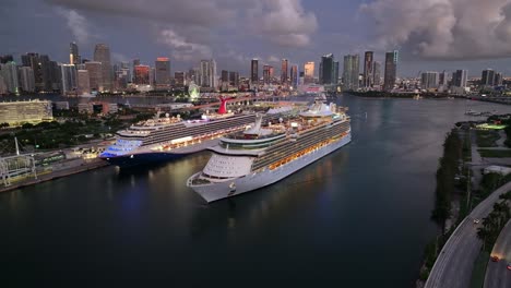 cruise ships docked at port of miami with the downtown miami skyline in the background. the scene shows vessels at dusk with city lights beginning to glow. aerial.