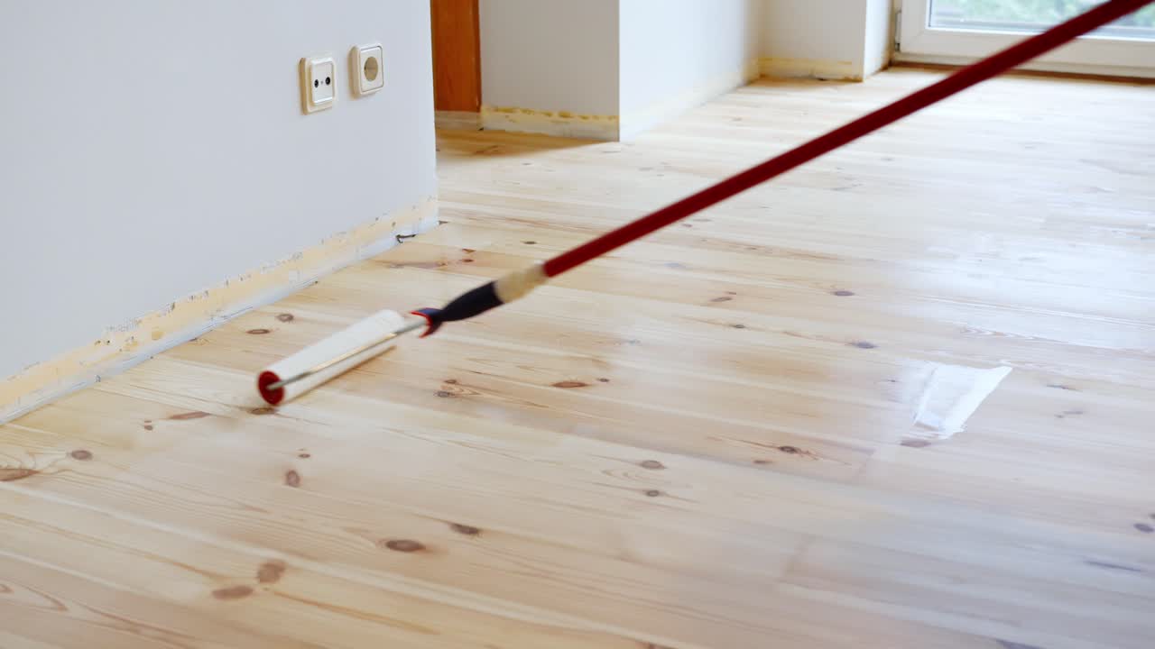 Roller applies glossy protective finish on sanded wooden floor in bright indoor room