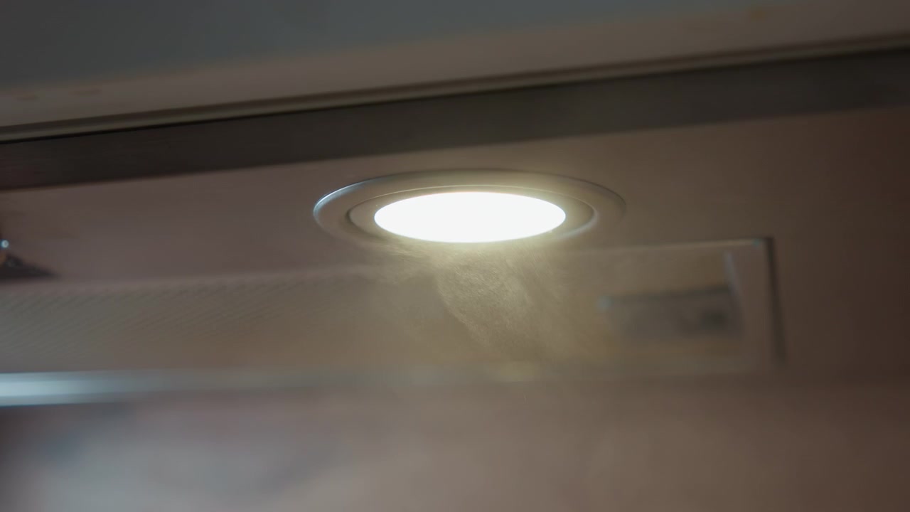 Nærbilde sett nedenfra av rundt innfelt lys under kjøkkenviften som lyser varmt, mens damp driver inn i lysstrålen og skaper en myk dis, med synlige partikler, teksturer av børstet metall og en rolig kjøkkenatmosfære