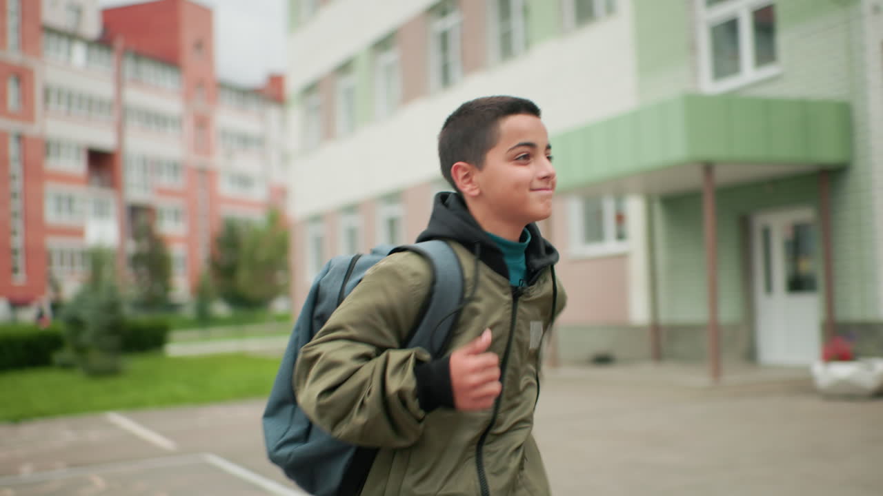 옆모습이에요. 어린 아이가 가방을 메고 계단을 오르면서 옆을 힐끗 보고 있어요. 흐릿한 주거 건물 배경에 결연함과 풋풋한 에너지가 느껴지는 표정이네요.