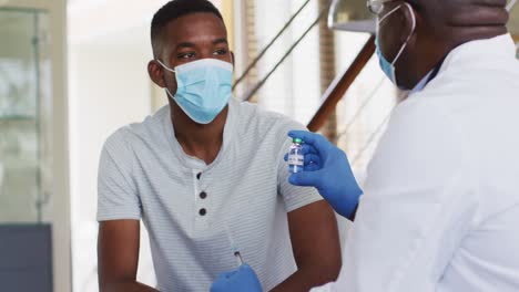 african american senior male doctor giving covid vaccine to male patient in home, wearing face masks