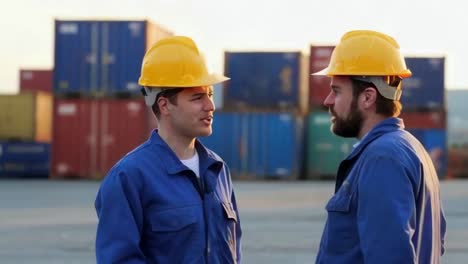 two men in blue work clothes and yellow helmet talking in port with containers in background.