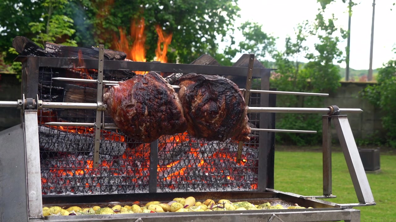Presunto de porco inteiro girando lentamente em um espeto de rotisserie sobre chamas de fogo a lenha ao ar livre na França. Batatas cozinham abaixo, recolhendo a gordura da carne em um banquete tradicional francês, similar ao mechoui