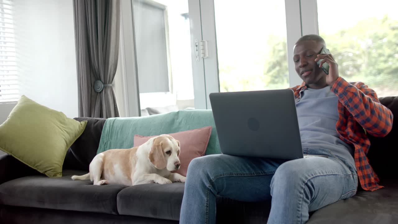 African american man talking on smartphone and using laptop at home, with his pet dog, slow motion