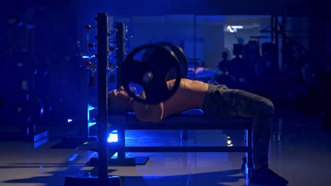 shirtless sportsman lifting weight. healthy man training with heavy barbell while lying on his back in the gym. side view. blue light filter.
