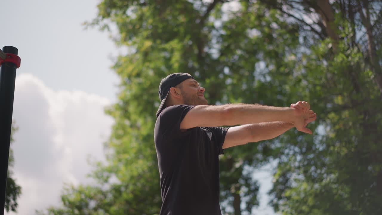 Man Performs Warmup Stretches Near Outdoor Refreshment Areas While Demonstrating Proper Form, Man Indicates And Extends His Arms As Part Of Training Warmup Beside Outdoor Cafes In Lush Park