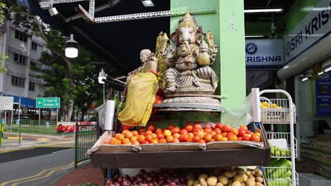 camera circles a vibrant produce display with tomatoes, potatoes, onions, and coconuts beneath a hindu deity statue at a street market in singapore