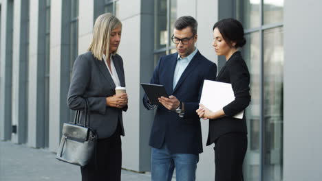 Hombre-De-Negocios-Con-Gafas-Y-Con-Tableta-En-Las-Manos-Contando-Y-Describiendo-Algún-Proyecto-De-Negocios-A-Dos-Mujeres-De-Negocios