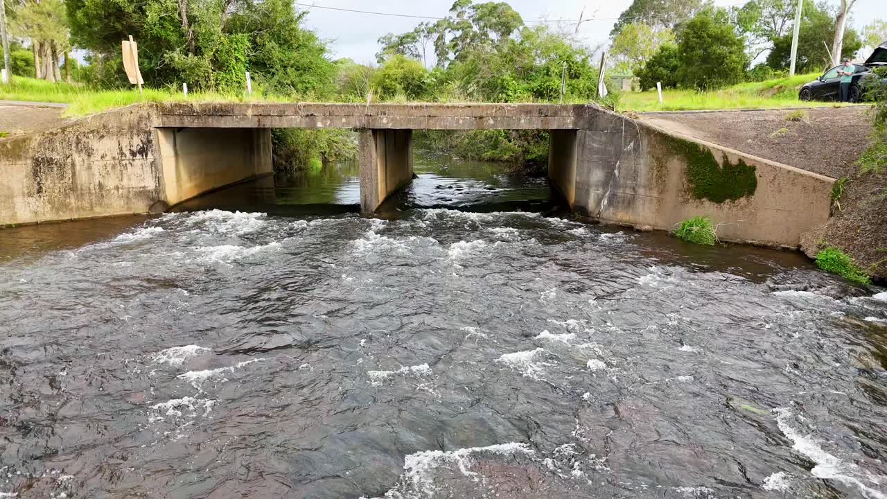 Steady stream flows beneath concrete bridge, surrounded by greenery, captured in daylight with static camera