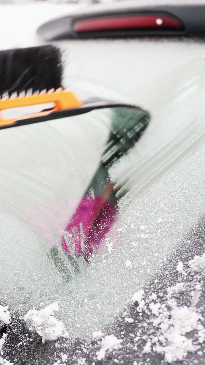Snow brush and ice scraper clear thick frost from a car rear windshield, revealing glass beneath as snowflakes fly. Vertical winter driving safety and cold weather b-roll.