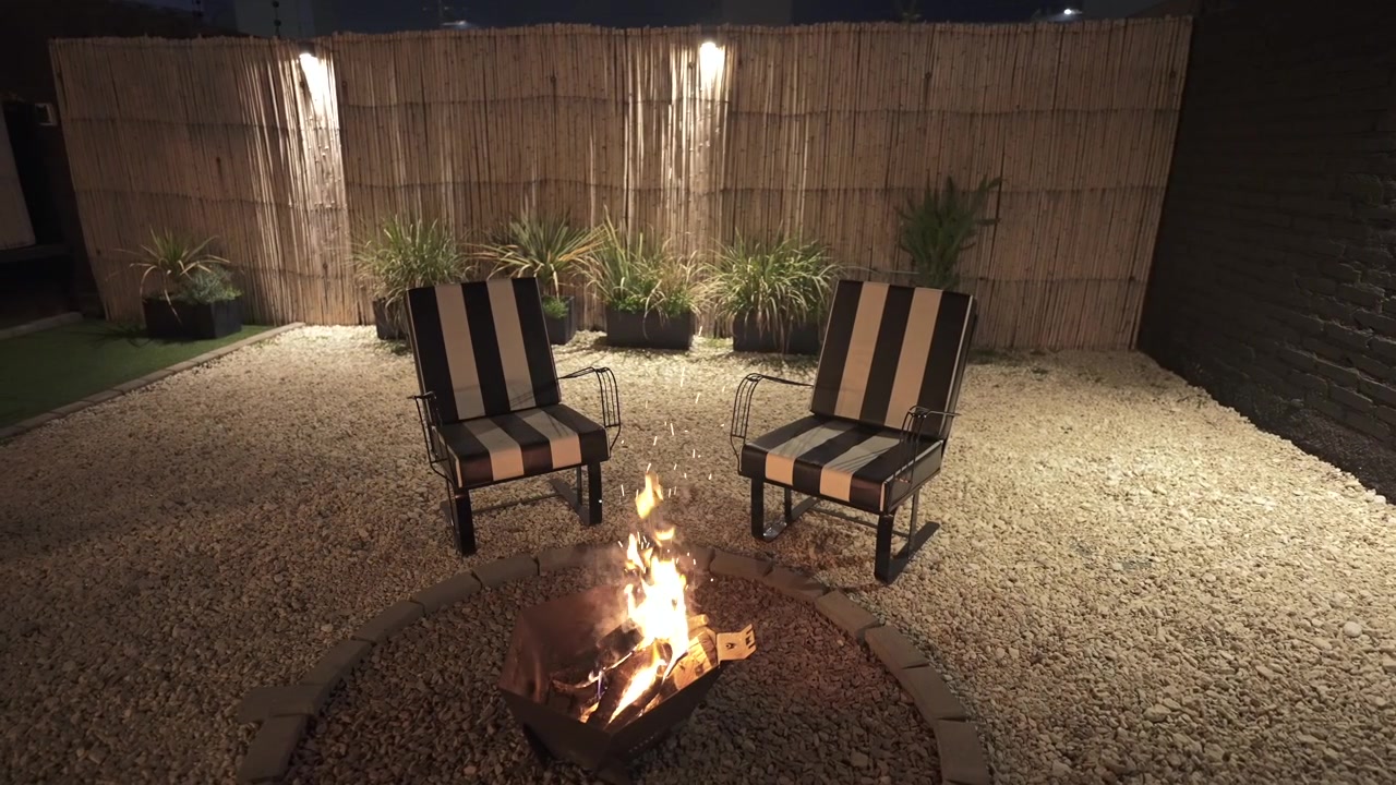 Scena notturna accogliente con un braciere ardente, due sedie a righe bianche e nere e piante ornamentali, ambientata in un patio ghiaioso con calda illuminazione ambientale a Mendoza, Argentina.