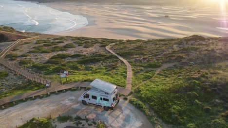 Drone-view-over-motor-home-car-standing-at-the-beach-during-sunset,-recorded-at-Bordeira-Portugal-hillside-coastline-overlooking-seascape-