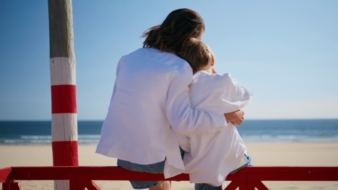 Happy mom child embracing sitting red fence by ocean waves.Mother hugging boy