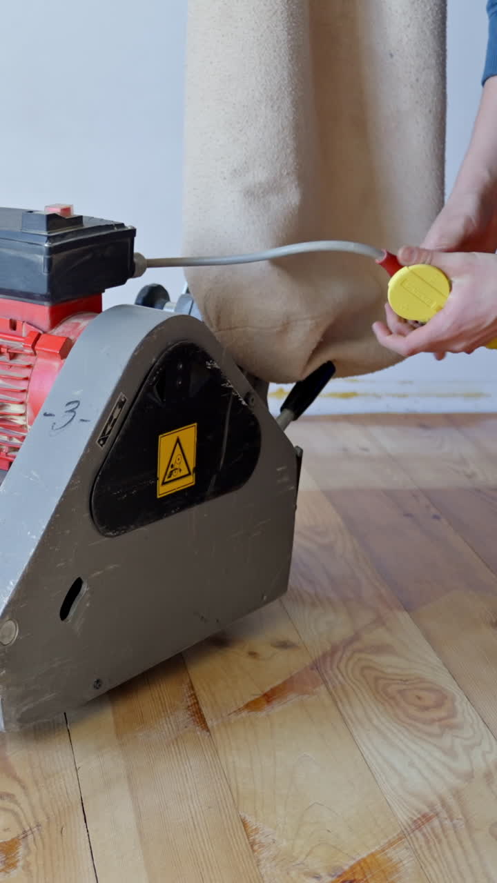 Vertical close-up shot of person’s hands connecting industrial floor sander to yellow electrical plug, ready for operation on pine sanded wooden floor, highlighting the preparation stage, static shot