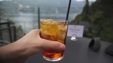 man rising glass of aperol spritz from table in restaurant by garda lake, italy