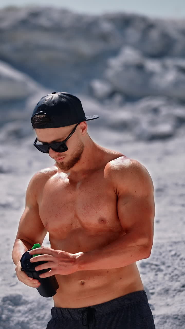 Topless sportsman drinking cold drink on natural background. Muscular man in black sunglasses drinks water after workout in the mountains. Vertical video