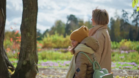 side view of mother walking with child while conversing through colourful autumn meadow blooming with flowers bordered by golden trees under overcast sky wearing warm jackets beanie scarf bag in park