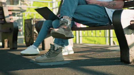 close-up of legs of two people seated outdoors on wooden benches, wearing casual sneakers and jeans, one holding a laptop while the other displays a tattooed hand, with sunlight casting shadows