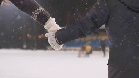 close up of middle aged mom in a warm sweater and her young daughter holding hands whilst ice skating at an ice-skating rink activity with snow falling down during winter break from hockey school