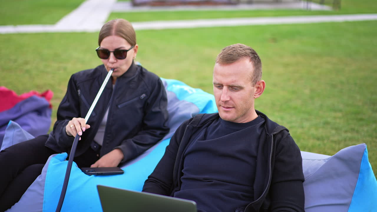 Mid-aged young couple in bean bag chairs resting outdoors. Male is focused on work on laptop and lady is smoking hookah. Summer time rest outdoors.