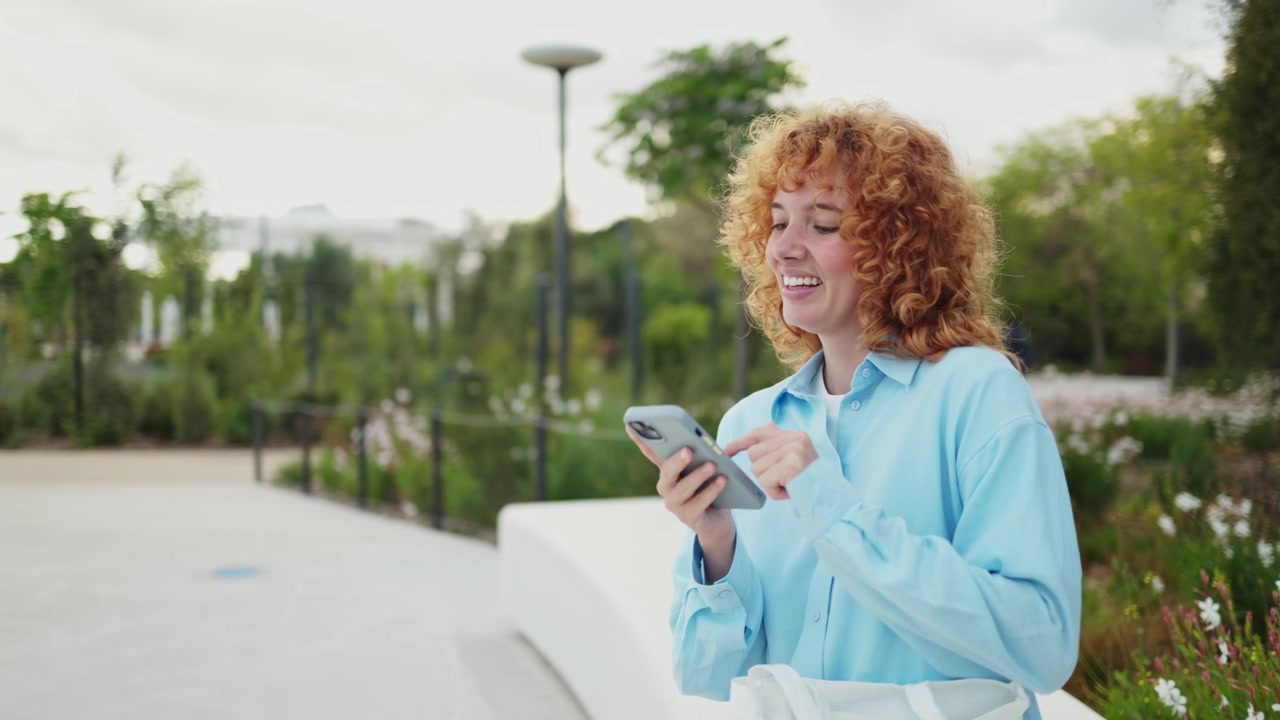 Happy Young Woman with Curly Red Hair Using Smartphone in Park
