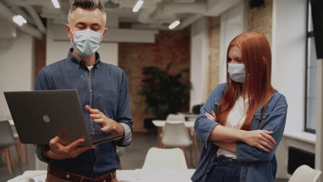 young business woman and man practice social distancing and work with a laptop wearing face masks in the office