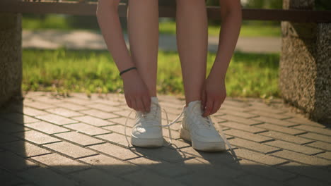 leg view of lady loosening shoelaces of her sneakers while seated on bench with bright sunlight casting soft shadows on pavement, with greenery in the background