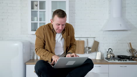 funny guy sitting on a kitchen table and typing on a laptop. young fellow studying on a laptop for long period. tired face of a freelancer working at home. remote job.