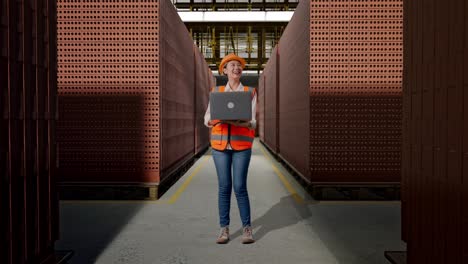 full body of asian female engineer with safety helmet working on a laptop and looking around while standing with red brick packed in stacks are stored