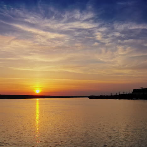 orange sun reflects in water surface and industrial factory nearby. beautiful evening background of setting sun on water. manufacturing territory with smoke from pipes near the river.