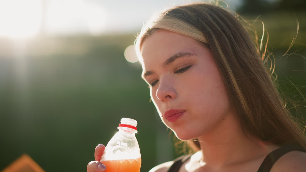 Lady sipping from a juice bottle outdoors, her focus drawn to something slightly blurred in the distance, sunlight creates a soft glow around her, with blurred background showing a lamp post