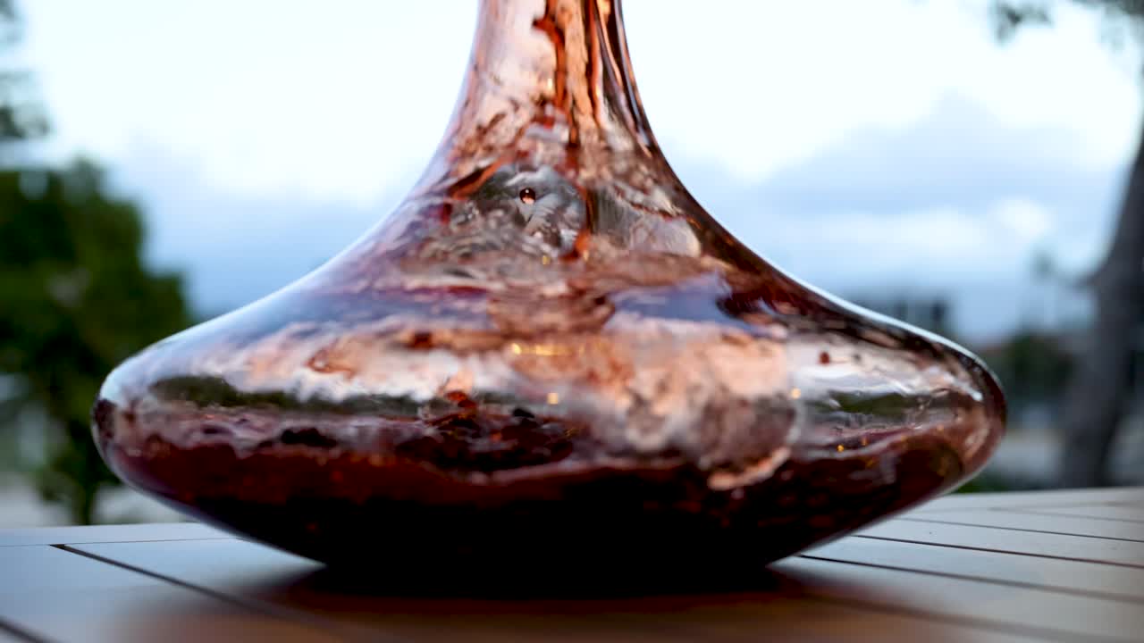 Wine swirls in a glass decanter on a wooden table, set against a natural outdoor backdrop