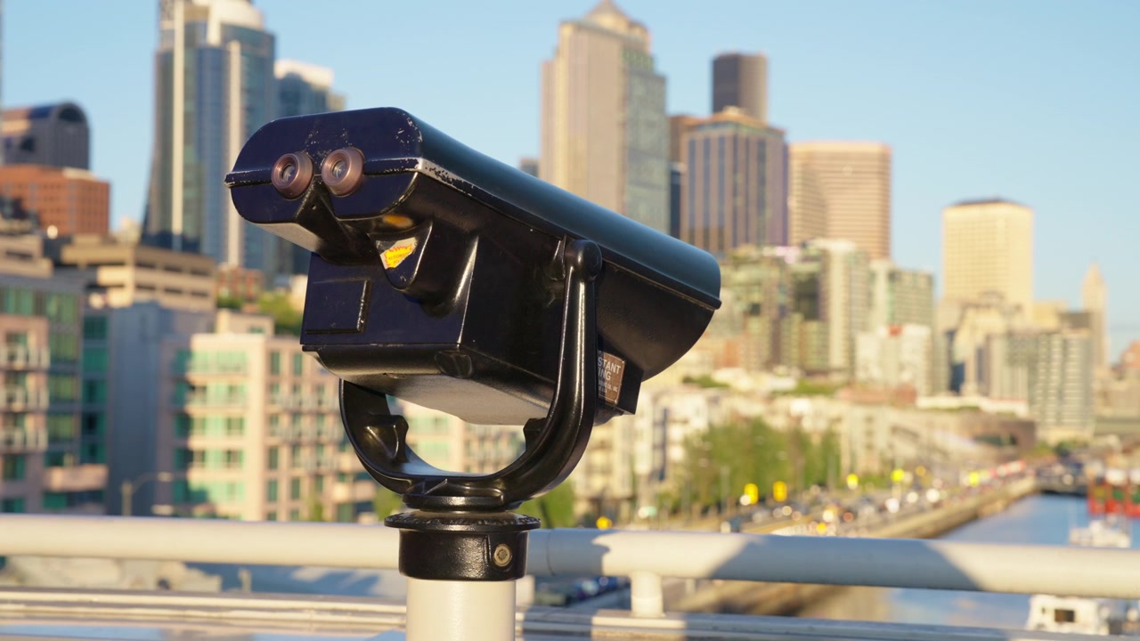 Camera orbits around a coin-operated viewfinder mounted on a railing, revealing the downtown Seattle skyline and city streets behind it in daylight, Orbit Shot
