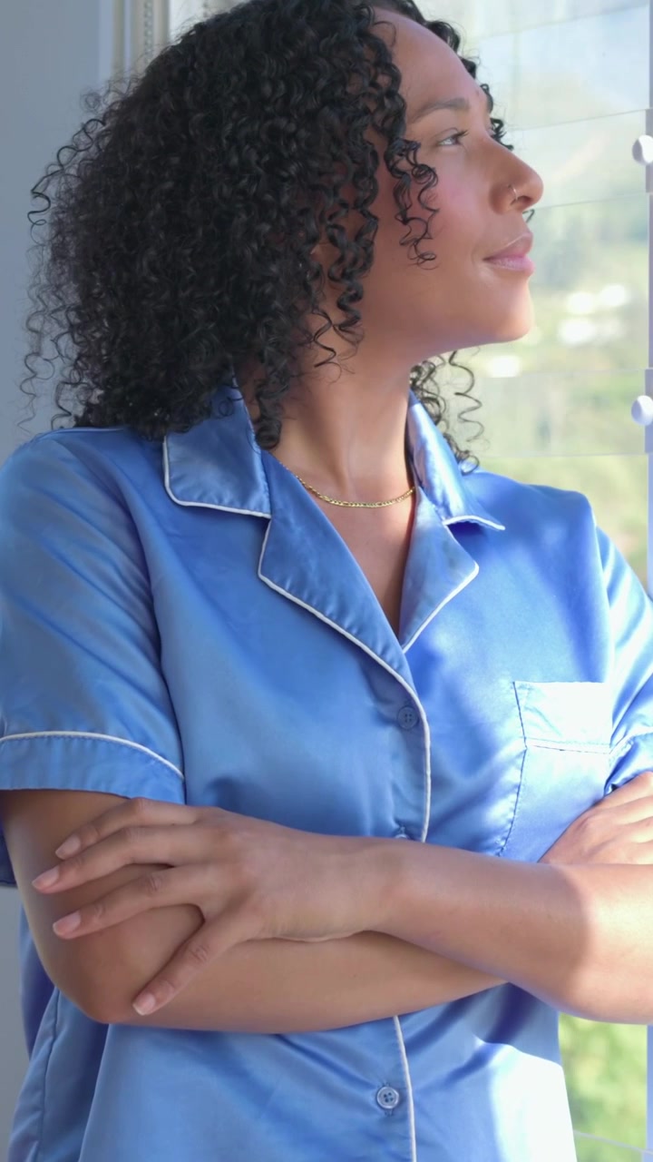 Video vertical: Mujer de pie en la ventana, con el sol, contemplando colinas, la cámara se acerca, lleva una blusa azul y una cadena de oro.