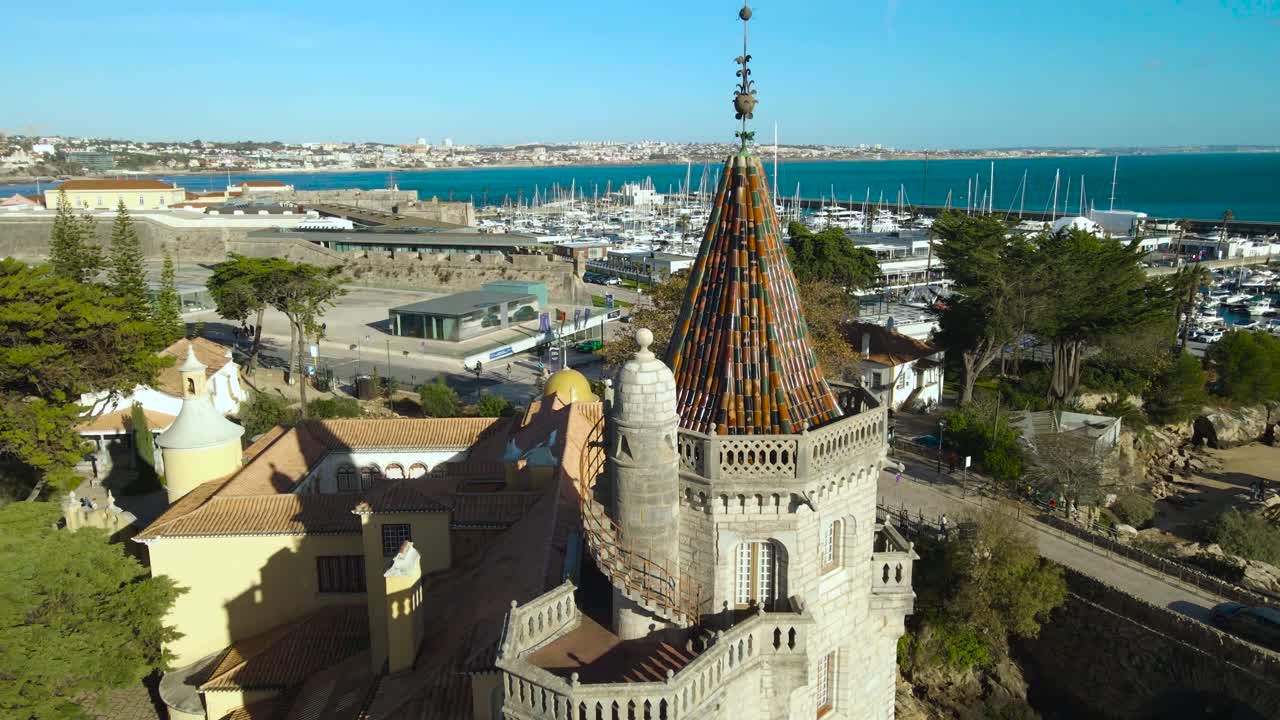 Aerial drone video descending in front of a museum and library tower in Cascais Estroil Portugal shoreline near Casa de Santa Maria viewing point during sunny day. Blue ocean water in the background