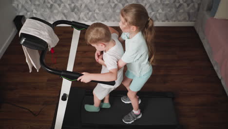 elder sister gently holds her younger brother from behind while he grips treadmill handle during shared indoor exercise, ensuring safety and balance