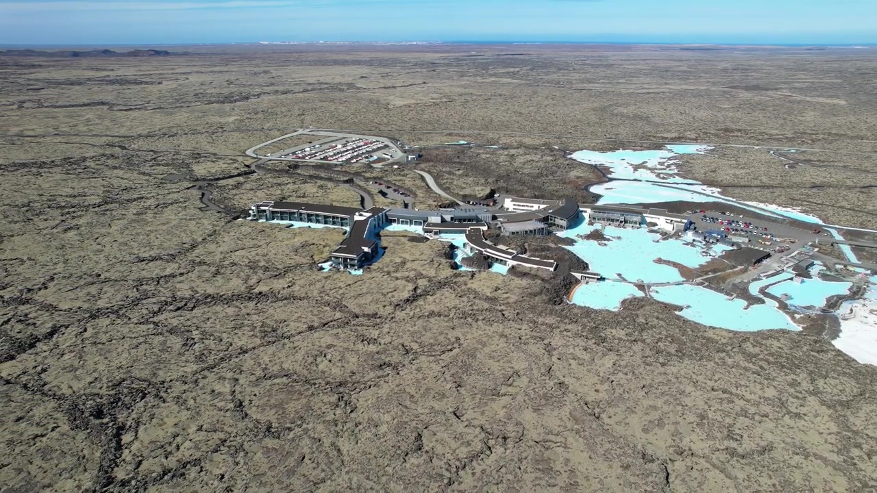 Vista aerea con drone della spa geotermica Laguna Blu, con vivide piscine di silice blu circondate da vasti campi di lava e terreno vulcanico sotto un cielo islandese limpido.