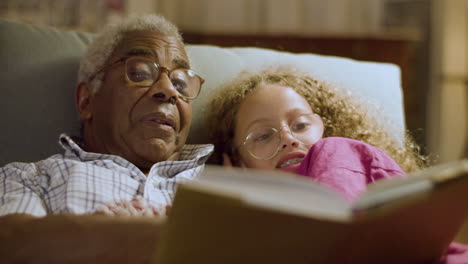 linda chica y su abuelo acostados en el sofá leyendo un libro juntos