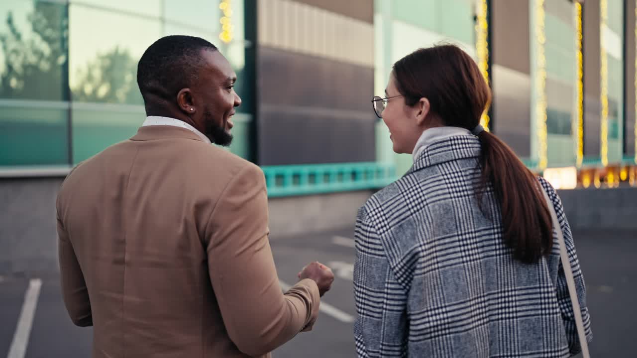 Rear view of a happy man with Black skin color and short haircut with a beard in a brown suit walks and talks with his colleague a brunette girl in a checkered gray coat during his business meeting in the city