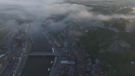 Wide-view-of-Dinant-city-next-to-maas-river-during-foggy-morning,-aerial