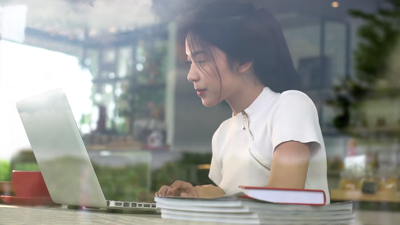 Portrait from a window of an Asian teenage girl working on a laptop in a cozy coffee shop on a bright sunny day, enjoying a relaxed atmosphere with warm natural light streaming into the café
